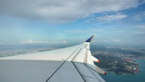 An aerial view from a plane window showing the aircraft wing, blue sky with clouds, and land and water below.