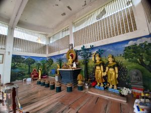 An interior view of a serene and colorful temple. The walls are adorned with a large mural depicting a lush landscape with trees and a body of water. In the center, there is a prominent altar with a golden Buddha statue flanked by two golden figures in traditional attire. The space features a mix of decorative items, including floral arrangements and offerings, on a wooden floor with a combination of light and dark tones. Natural light filters through the slatted windows above, creating a peaceful ambiance.