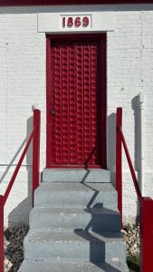 A red steel door set into a white wall with gray stairs and red hand rails. The numbers 1869 are written over the door.