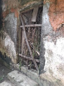 A rustic wall with peeling paint and damp patches features a small barred window stuffed with dry branches.