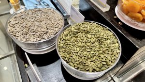 Two glass bowls at a buffet are filled with shelled sunflower seeds and green pumpkin seeds for toppings or snacks.