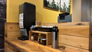 A wooden counter with things needed by coffee shop customers. Eating utensil dispenser, napkins, sugar packets, creamer bottler, etc.