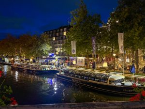 Illuminated canal boats docked along the water at night, with signs for “Blue Boat” and “We Are Hiring” visible. Trees and city buildings glow with lights in the background, while people walk and cycle along the lively street.