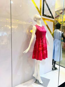 A white female mannequin models a bright pink tiered dress and a necklace in a brightly lit store window with geometric gold and black accents.