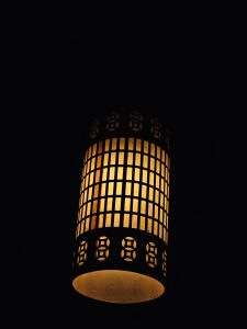 A glowing cylindrical hanging lamp with patterned cutouts lights up the dark, captured indoors in Kozhikode, Kerala. Its warm orange tone creates a cozy atmosphere. 
