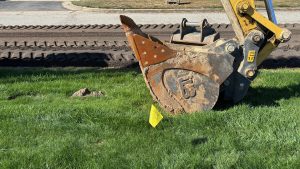 The bucket from a back hoe resting on grass, a plowed road in the background.