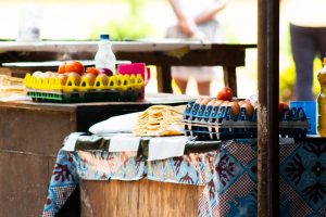 A wooden stall displays various food items including a green plastic tray filled with eggs, a stack of tortillas, and a few tomatoes. A bottle of water and a colorful cup can also be seen on the table, which is covered with a patterned cloth. In the background, a blurred figure stands, suggesting a bustling market environment.
