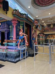 The entrance of a richly decorated Hindu temple features vibrant blue walls, multi-colored pillars, and statues of deities in dynamic poses, with a polished marble floor and railings in the foreground.