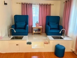 A pedicure station with two tall blue chairs, white sinks, and pink curtains.