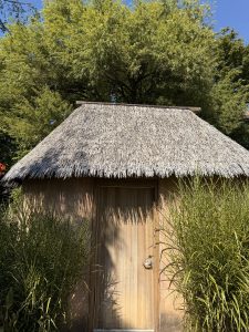 A small wooden hut with a thatched roof, surrounded by tall green grass, photographed inside the Oregon Zoo in Portland, Oregon. 