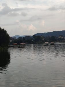 A serene lake with calm waters reflecting the sky and hills, dotted with canopy boats under a cloudy sky.