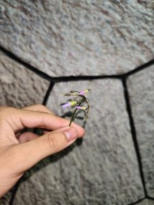 A hand holds a delicate flower with thin green stems and small purple petals, positioned in front of a textured stone background.