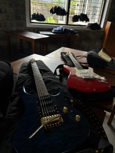 A close-up view of two electric guitars resting on a black guitar bag, with a wooden table in the background. One guitar is dark blue with gold hardware, and the other is bright red with white accents. There's a window in the background with clothing hanging, and bags placed on the table. The walls are made of stone, giving the room an industrial feel.