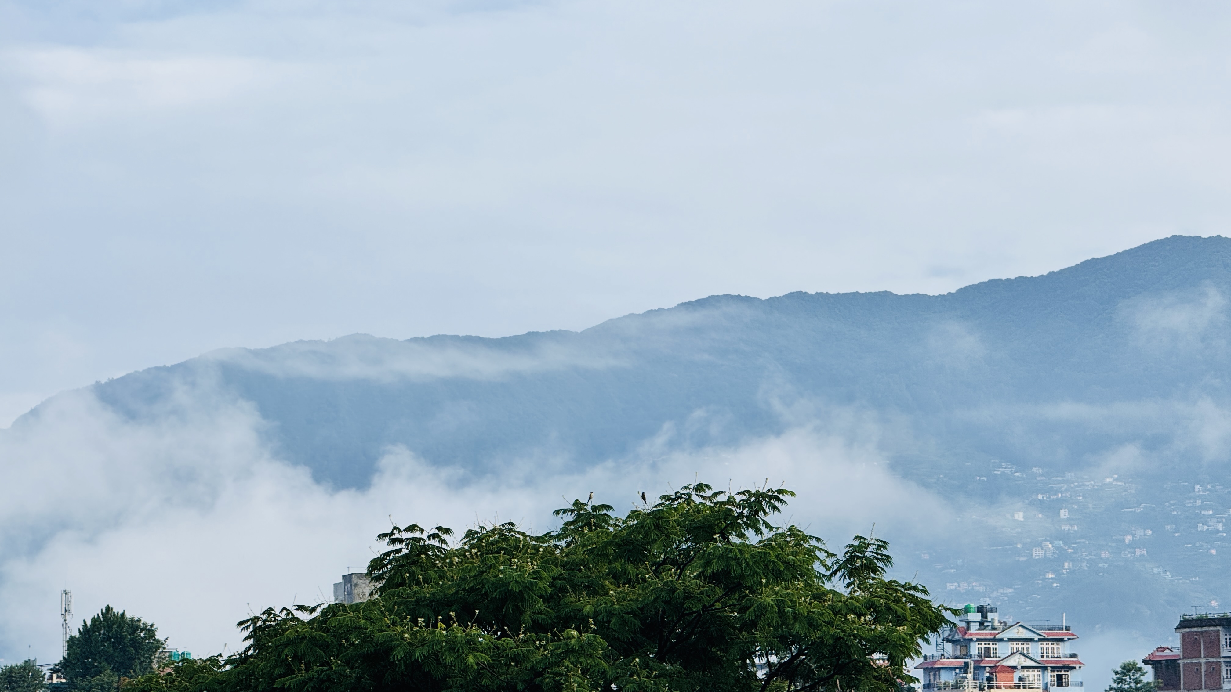 A panoramic view of the misty mountains in the Budhanilkantha area of Kathmandu, with low-hanging clouds partially obscuring the peaks under a cloudy sky.