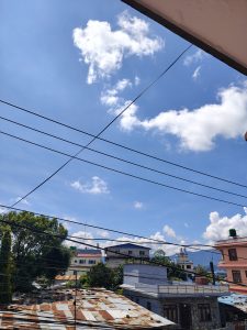 A clear blue sky filled with fluffy white clouds, intersected by multiple power lines. Below, the rooftops of several buildings are visible, with a mix of textures and colors, including a rusty metal roof. 
