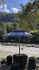 A blue umbrella stands upright on a sandy riverbank near a rushing stream. Surrounding the umbrella are several wooden stools. 