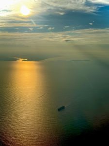 A cargo ship sails across a calm sea at sunset, with golden sunlight reflecting off the water and islands faintly visible on the horizon under a dramatic sky with scattered clouds.