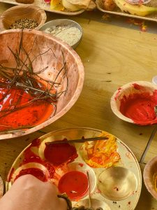 An overhead view of various ritual items, including a large copper bowl with red liquid and pine needles, small bowls of rice and seeds, and a brass palette with red and orange pastes.