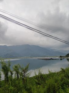 

A calm Fewa lake surrounded by lush greenery, with distant mountains partly in clouds.