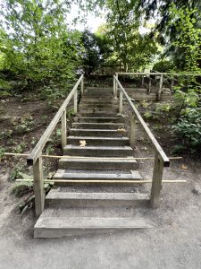 A rustic, closed wooden staircase with railings leads through a shaded forest area within the Japanese Garden in Portland, surrounded by lush green foliage and fallen leaves. 
