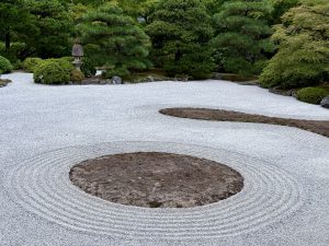 A wide view of a raked gravel garden with circular patterns and islands of moss. Pine trees and bushes surround the Zen space. Taken at Portland Japanese Garden. 