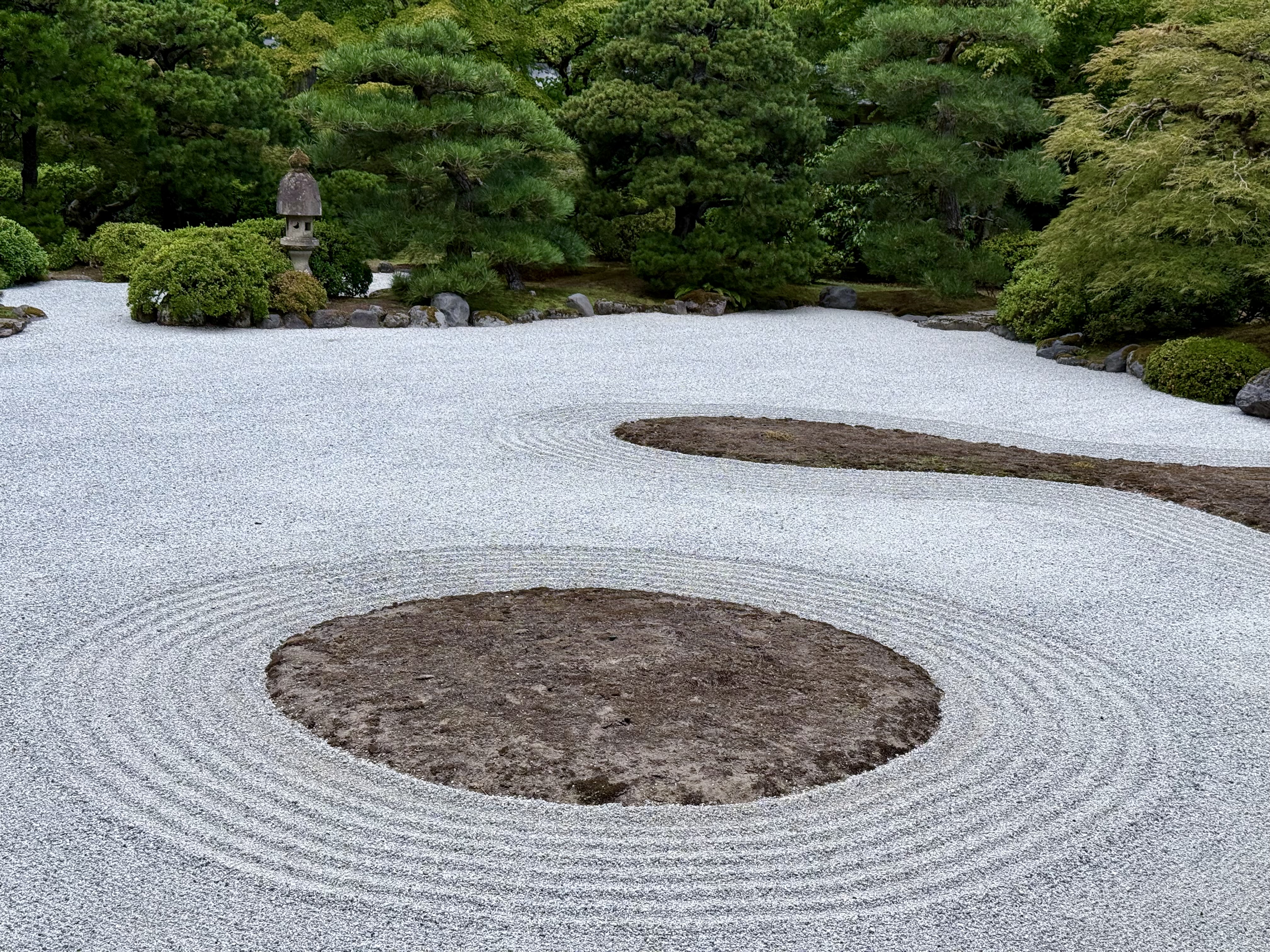A wide view of a raked gravel garden with circular patterns and islands of moss. Pine trees and bushes surround the Zen space. Taken at Portland Japanese Garden. 