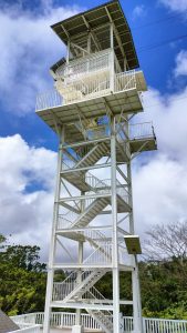 A white observation tower with a spiral staircase, topped by a platform, set against blue skies and greenery.