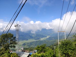 A tranquil mountain landscape under a bright blue sky with white clouds. A cell tower and power lines stand among trees, leading toward green mountain layers, a winding river, and farmland in the valley below.