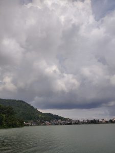 Large expanse of water, a settlement in the distance, the left is edged by a hillside covered in green vegetation, cloud grey sky looms above.