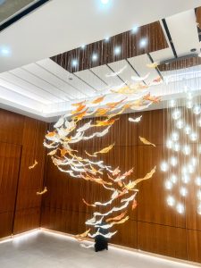 A large modern light installation hangs from a high ceiling, featuring a swirling school of orange and white glass fish suspended by thin wires above a polished wood-paneled wall.