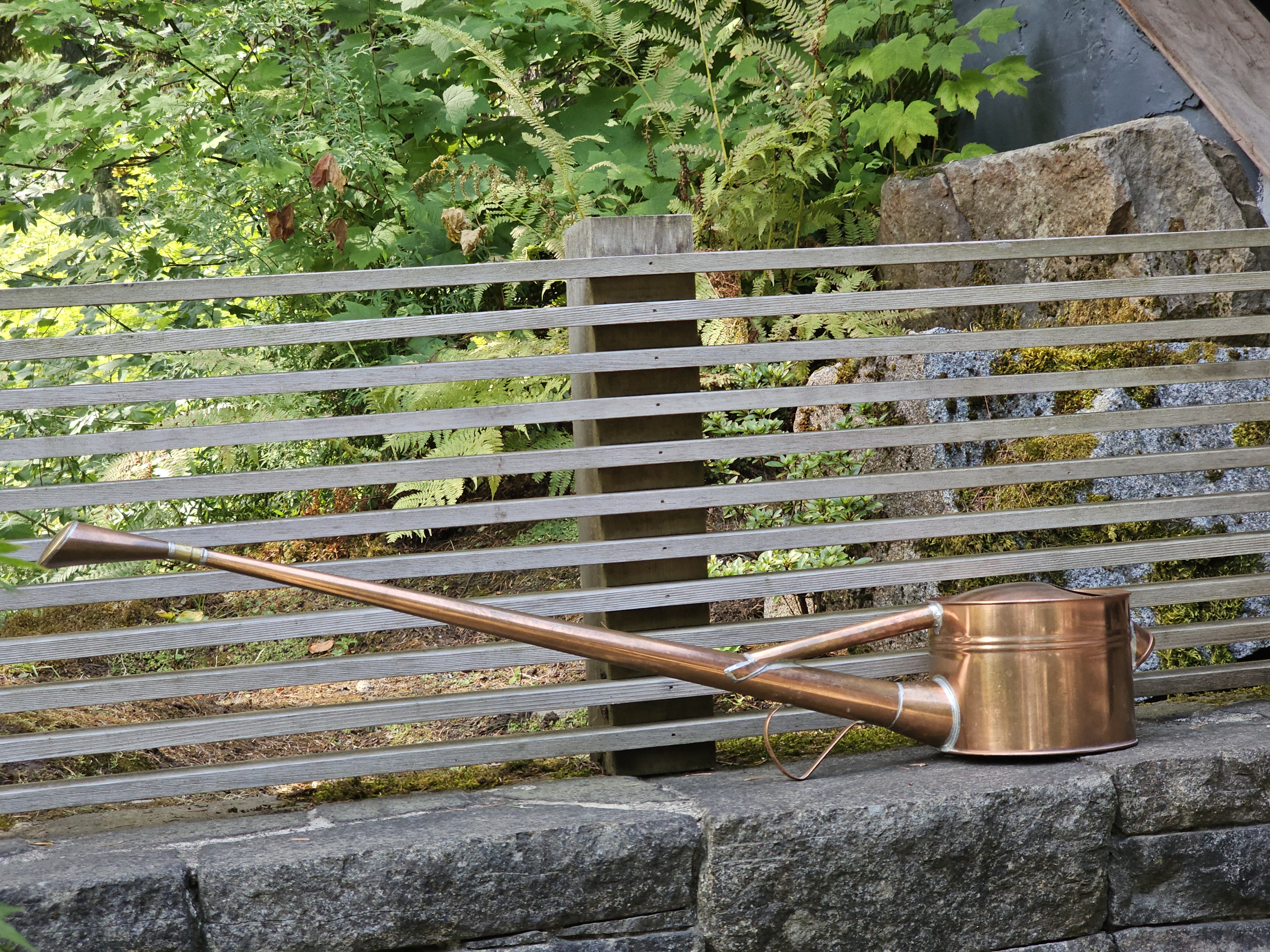 A copper bonsai watering can with a long spout rests on a stone wall in a peaceful garden. Green foliage and a wooden fence complete the background. Captured at the Portland Japanese Garden, Oregon.