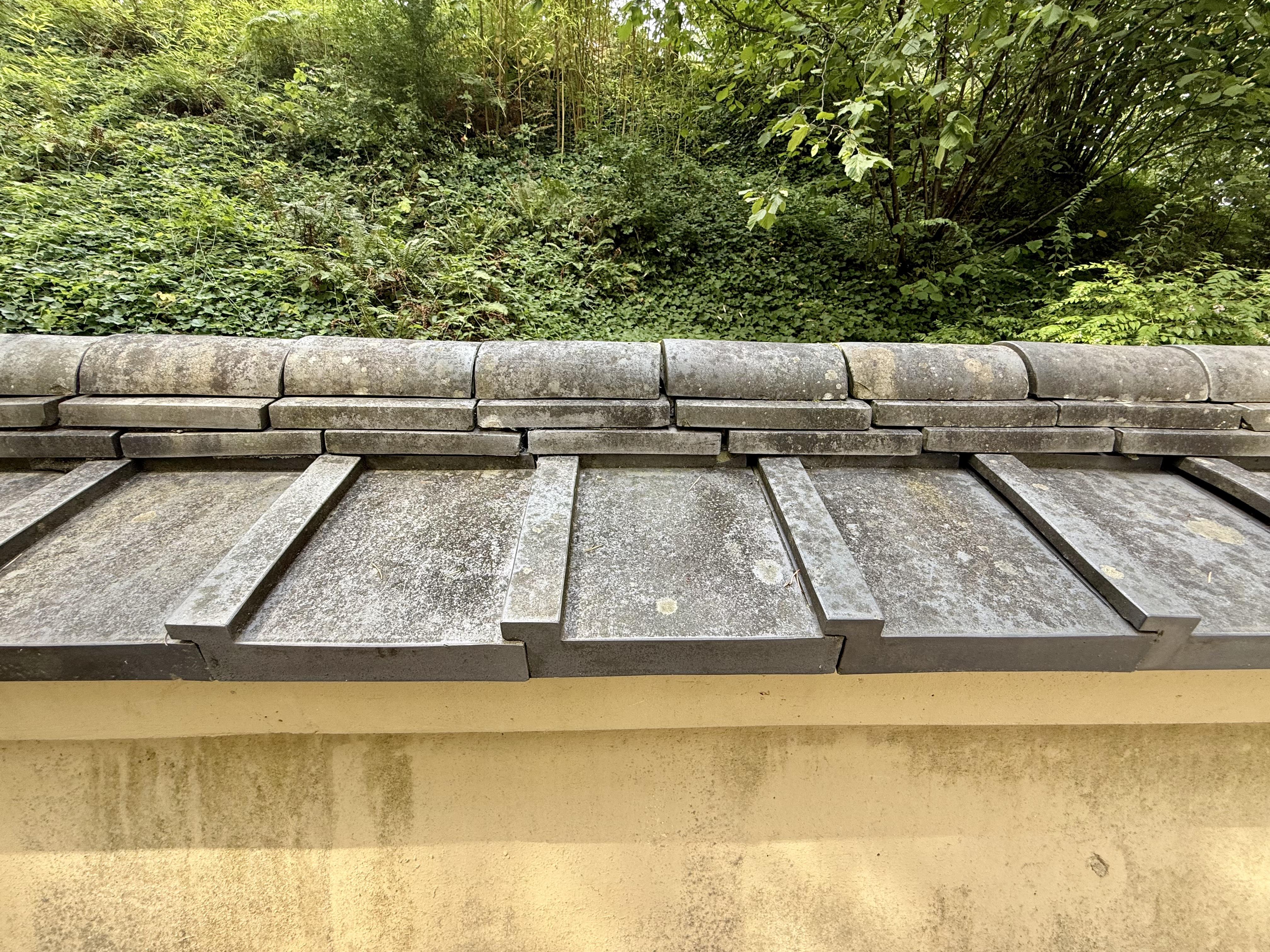 Close-up of a traditional Japanese-style wall with layered roof tiles in the Portland Japanese Garden. Moss and plants grow naturally behind the wall. 