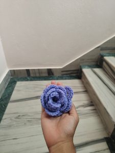 A hand holding a handmade crocheted flower in shades of light purple, with a textured, layered appearance. 