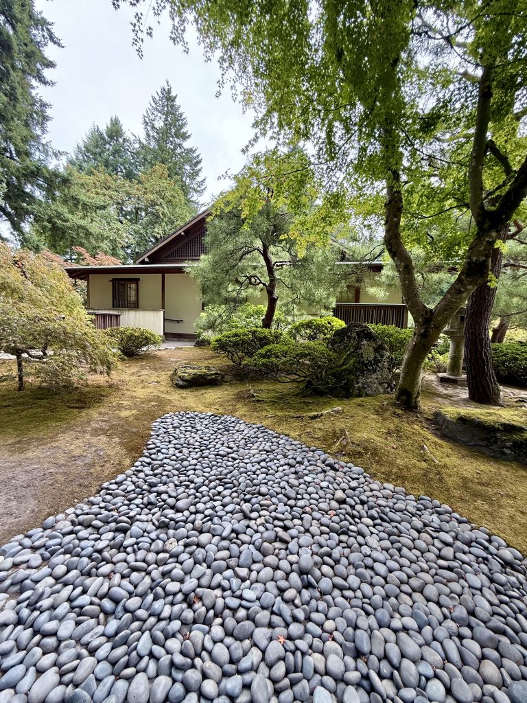 A traditional Japanese-style building behind a curved pebble stone path, surrounded by trees and moss. Photo taken at Portland Japanese Garden, Oregon, USA.