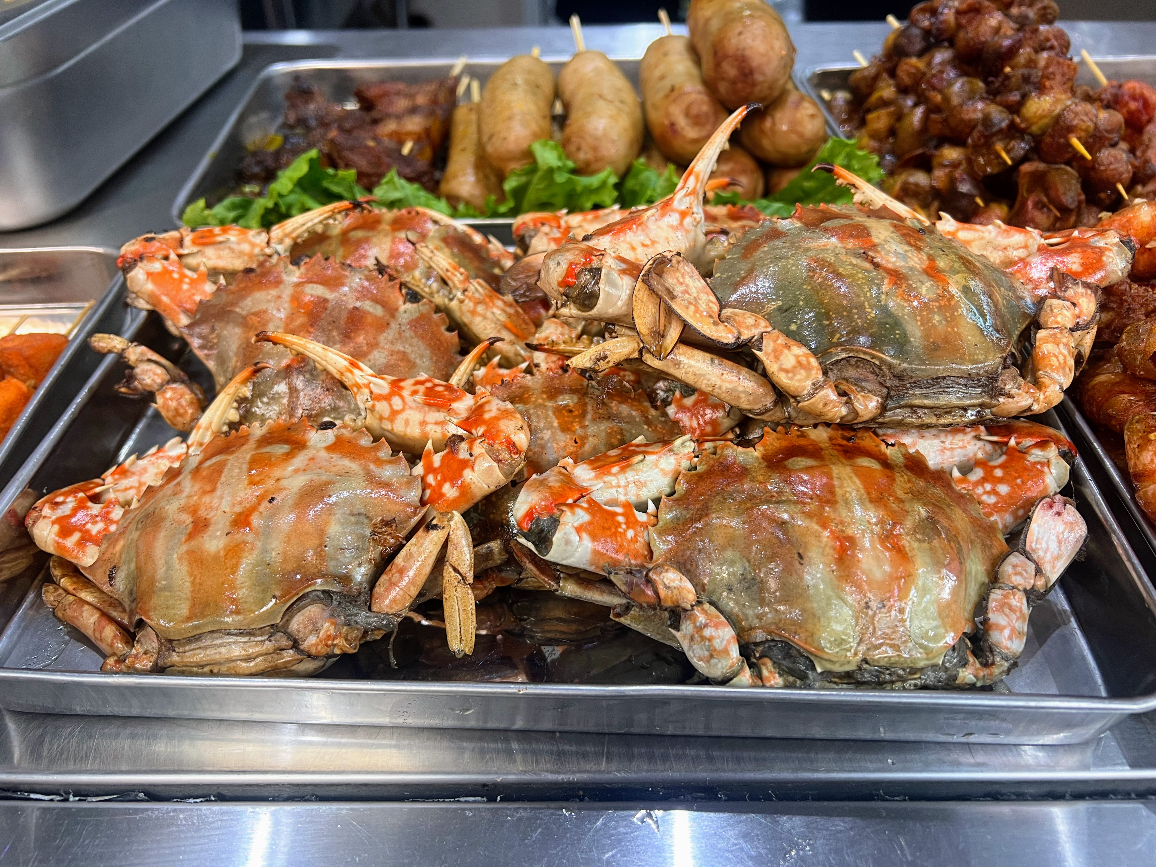 A close-up view of a metal tray filled with cooked red and brown crabs, alongside skewers of potatoes and other snacks.