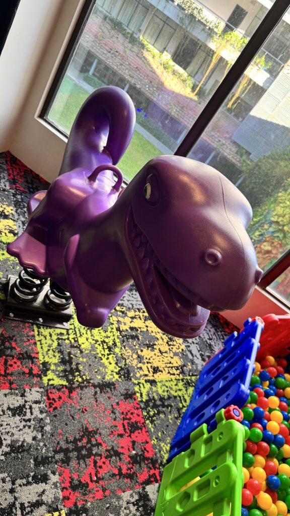 A bright purple, spring-mounted rocking toy shaped like a smiling dinosaur sits in a children’s play area with a colorful carpet.