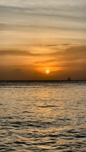 A sailboat is silhouetted on the horizon of the calm ocean as the sun sets.