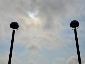 Two lampposts stand against a bright, cloudy sky with the sun partially obscured, creating a serene and minimalist scene. Captured from World Forestry Centre, Washington Park, Portland.