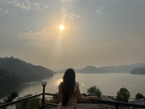 A girl standing on a balcony railing, looking at the sun setting over a large lake surrounded by rolling hills.