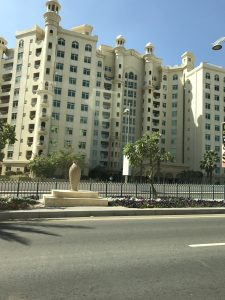 A large, ornate white building with many windows and balconies, seen from a street with manicured bushes and trees.