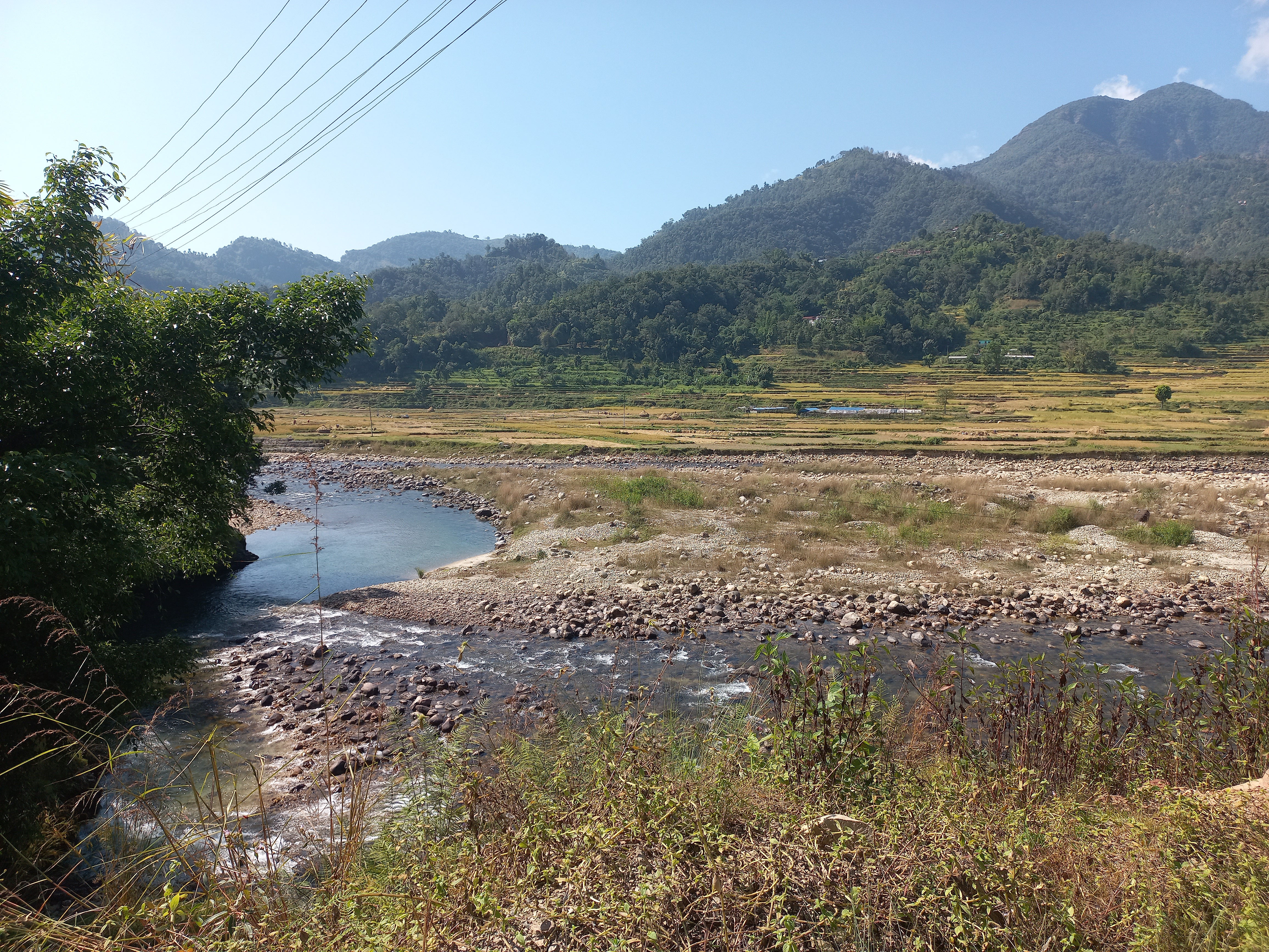 A serene landscape with a winding rocky river, lush greenery, terraced fields, and distant mountains under a clear blue sky, with power lines overhead.