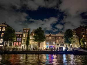 A row of traditional canal houses at night, their windows softly lit and reflecting colorful lights onto the rippling water below. Trees line the waterfront, while a partly cloudy sky adds depth to the evening scene.
