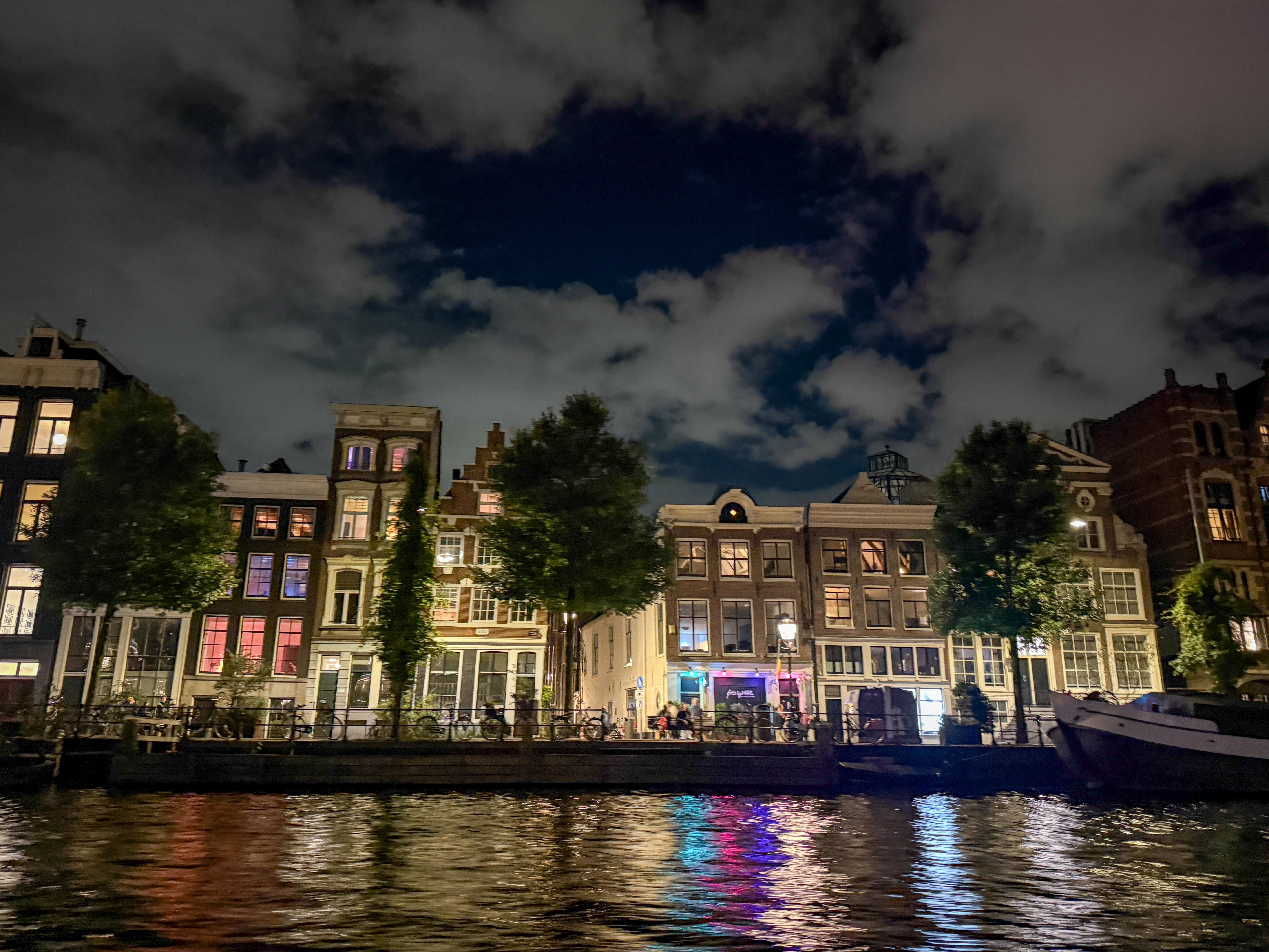 A row of traditional canal houses at night, their windows softly lit and reflecting colorful lights onto the rippling water below. Trees line the waterfront, while a partly cloudy sky adds depth to the evening scene.