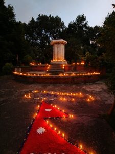 A tranquil evening scene of a Nepal monument on a circular stone platform, surrounded by glowing oil lamps and a flag-inspired ground design, with lush greenery in the twilight backdrop.
