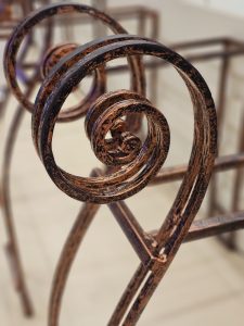 Artistic close-up of a rusted spiral iron frame used to control the queue, showing texture and shape. The background is softly blurred. Captured in Kozhikode. 