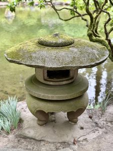 A moss-covered stone lantern stands beside a pond under a leafy tree in the Japanese Garden, Portland, representing traditional Japanese design and peaceful scenery. 