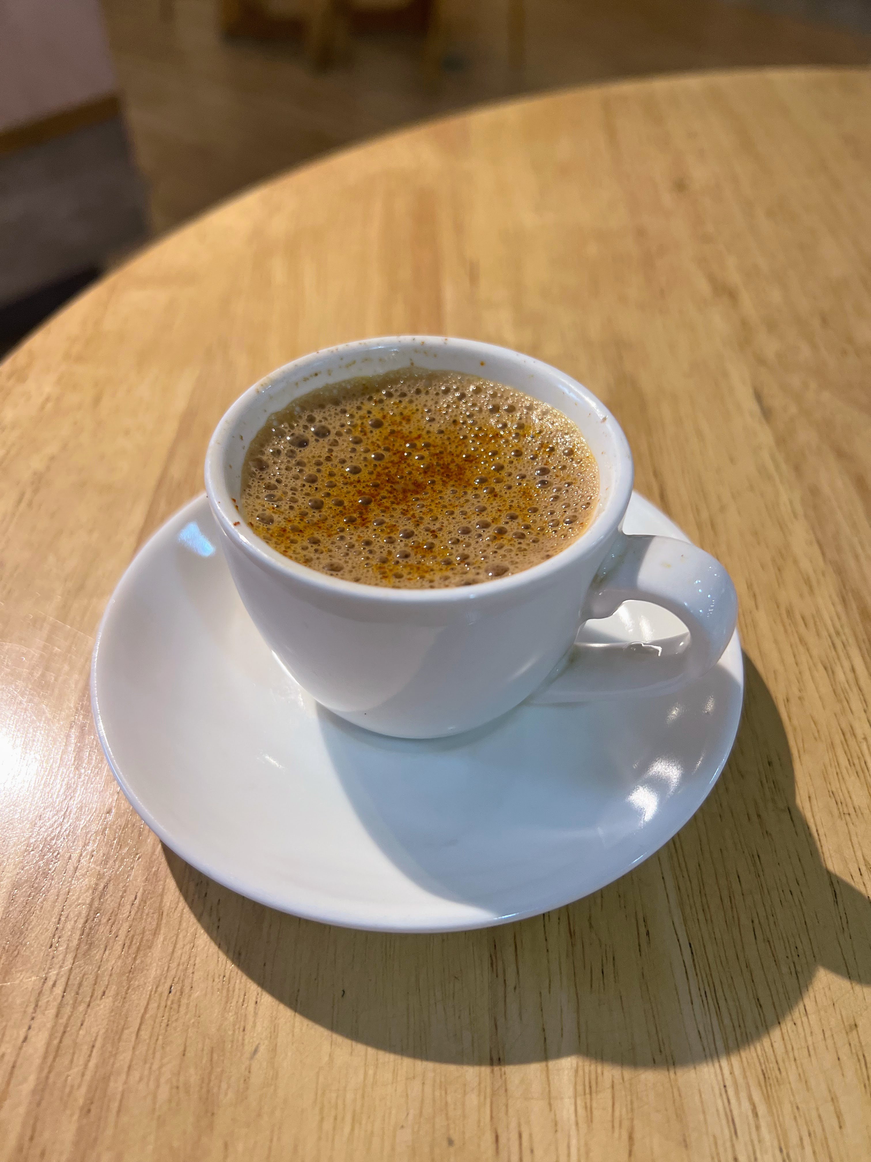 A white cup and saucer holding a hot, frothy brown beverage - Zafaran Tea, sits on a wooden table.