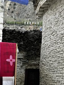 A narrow, rocky alleyway in a village with stone buildings. A red blanket with a white flower design hangs on a stand to the left, and the word "MARPHA" is spelled out on a hillside in the background.