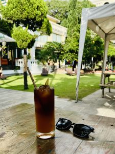 A tall glass of iced coffee or tea with a straw sits next to a pair of sunglasses on an outdoor wooden table, with a green lawn and building in the background.