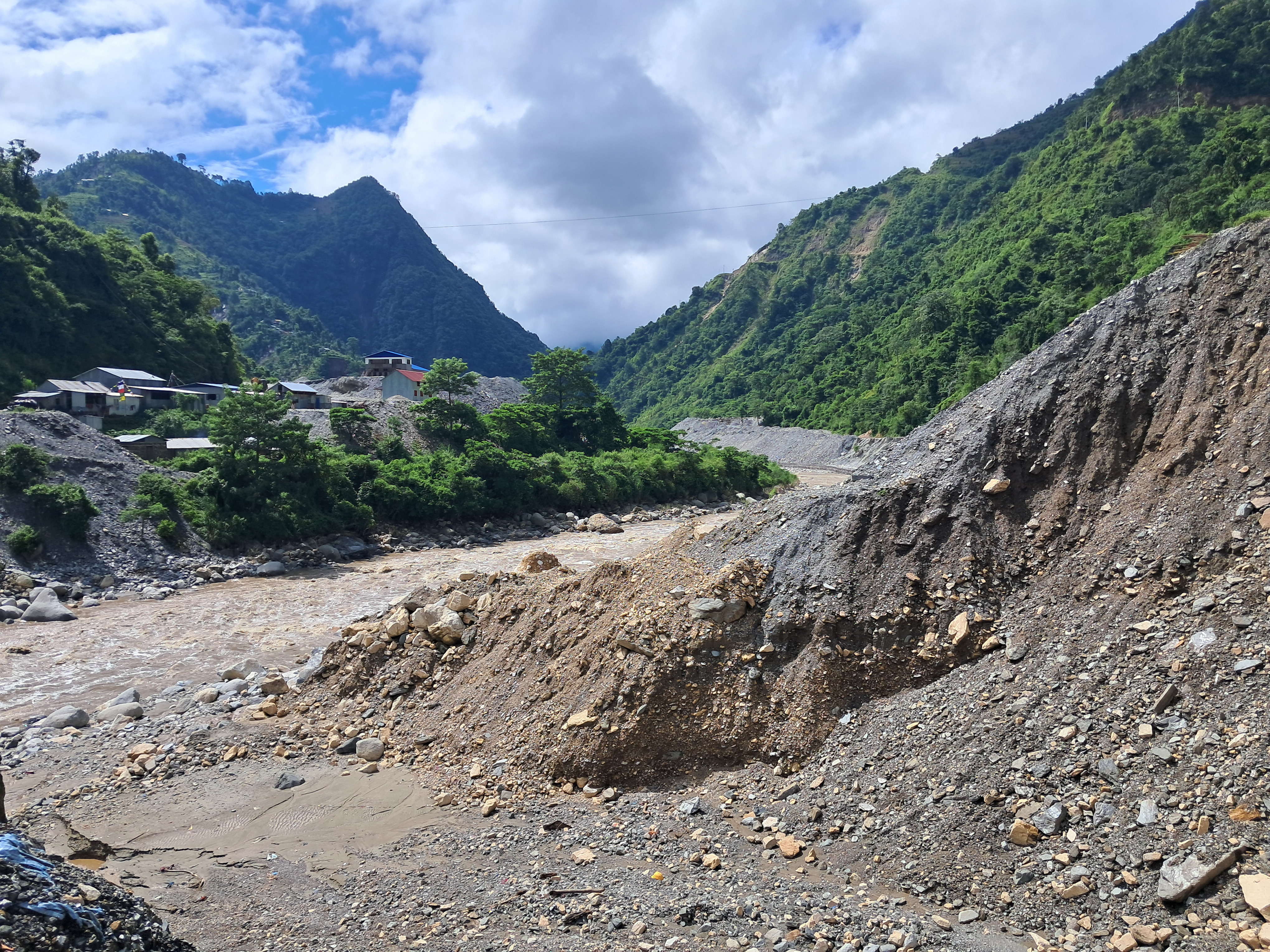 A wide, brown river flows between steep, rocky banks and lush green hills. A few buildings are visible on the left side of the river, and the sky above is partly cloudy.